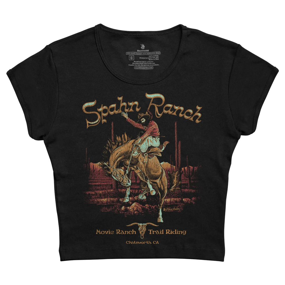 Black short-sleeved t-shirt featuring a vintage-style illustration of a cowboy riding a bucking horse with ’spahn ranch’ and ’movie ranch trail riding chatsworth ca’ printed in gold lettering.