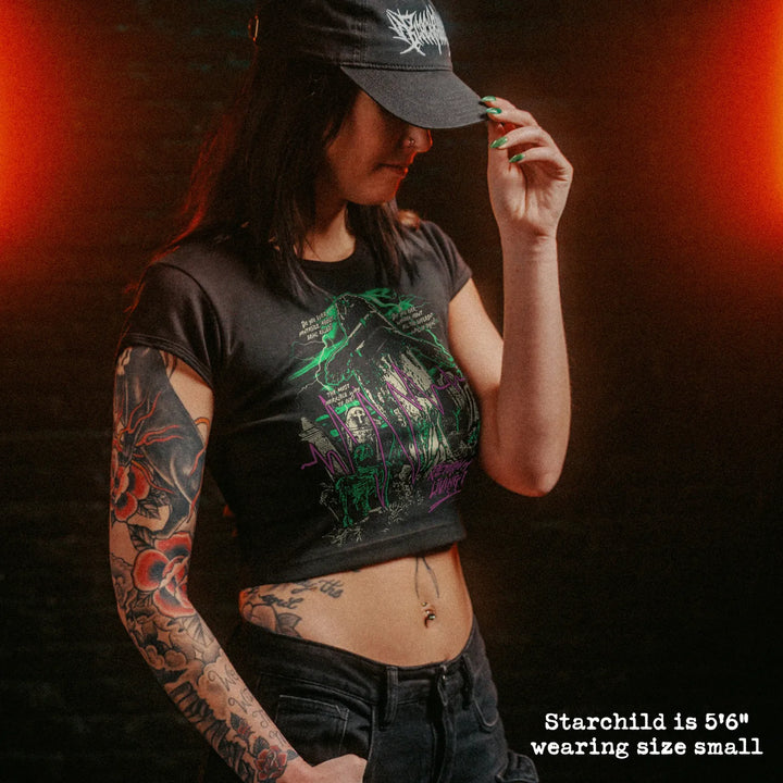 A woman wearing a black graphic crop top t-shirt with 'Return of the Living Dead' text and artwork, paired with a black cap and jeans, set against a dark brick wall with dramatic orange lighting.