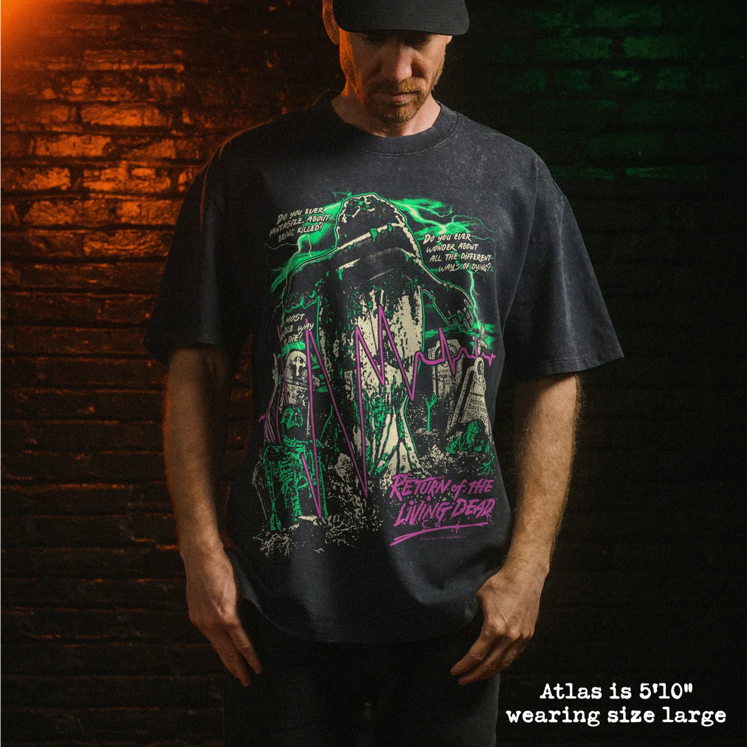 A man wearing a dark charcoal graphic t-shirt with a green and purple horror-themed print, standing against a dark brick wall with dramatic orange and green lighting.