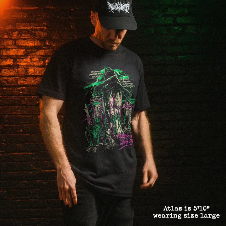 A man wearing a black short-sleeve graphic t-shirt and a black baseball cap with a white logo, standing against a dark brick wall with dramatic orange and green lighting.
