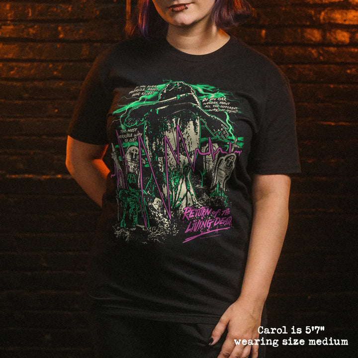 A woman wearing a black graphic t-shirt with a green and purple horror-themed print, standing against a dark brick wall background.