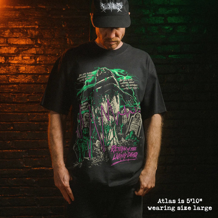 A man wearing a black graphic t-shirt and a black baseball cap stands against a dark brick wall lit with orange and green lighting. The t-shirt features a detailed graphic of green, purple, and white, with the text 'Return of the Living Dead'.