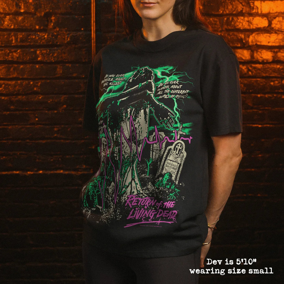 A woman wearing a black graphic t-shirt with a gothic zombie design, standing in front of a dark brick wall. The graphic features a zombie, tombstones, and green and purple text reading 'Return of the Living Dead'.