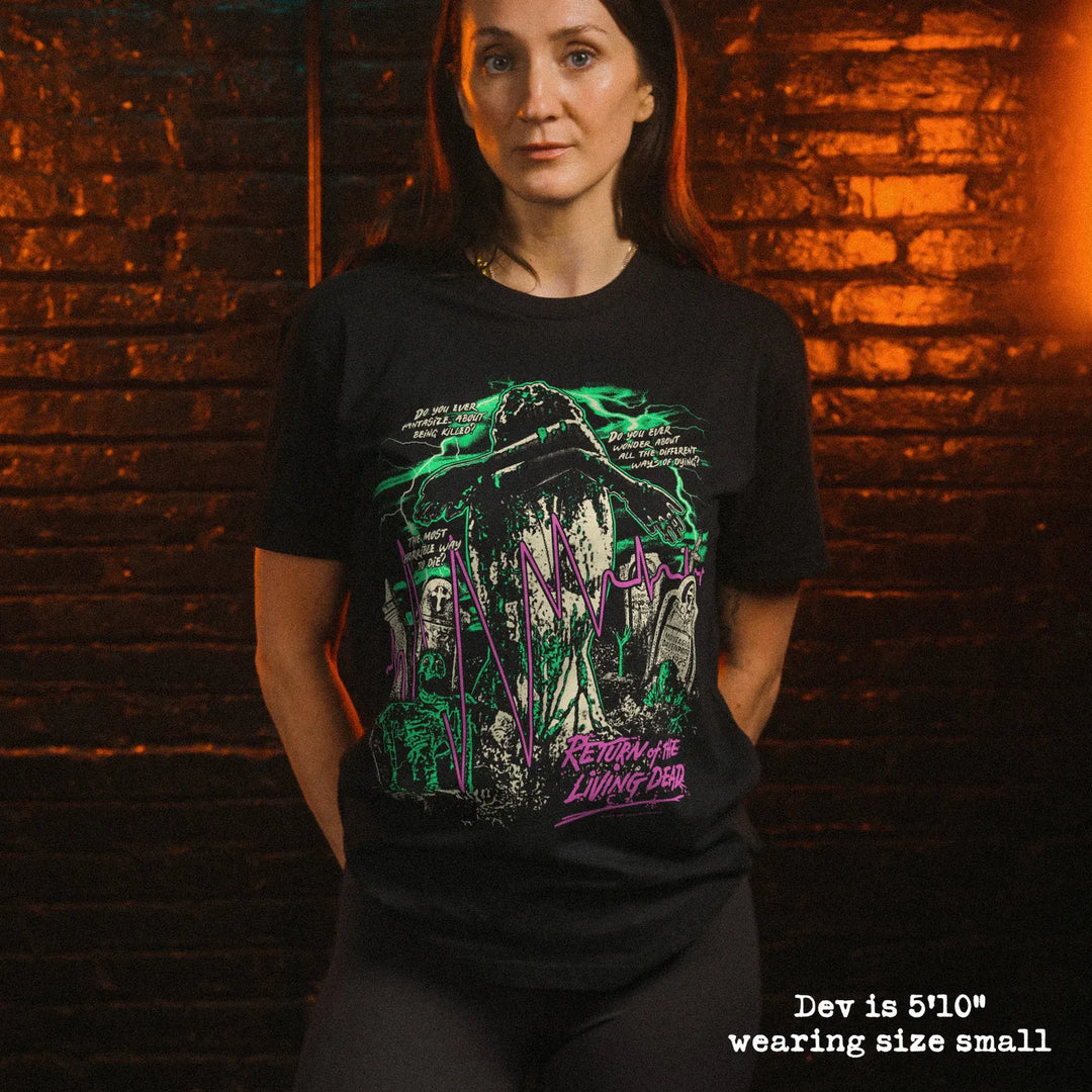 A woman wearing a black graphic t-shirt with a green and purple horror-themed print, standing in front of a dark brick wall with warm lighting. Text in the bottom right corner reads 'Dev is 5'10" wearing size small'.