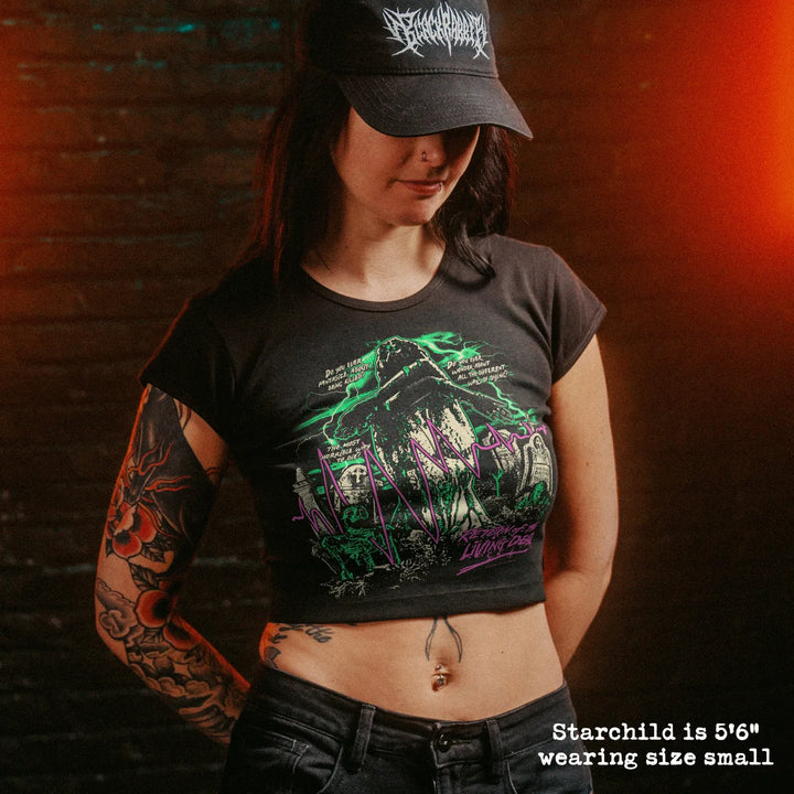 A woman wearing a black crop top t-shirt with a green and purple graphic, set against a dark brick wall with dramatic orange lighting.