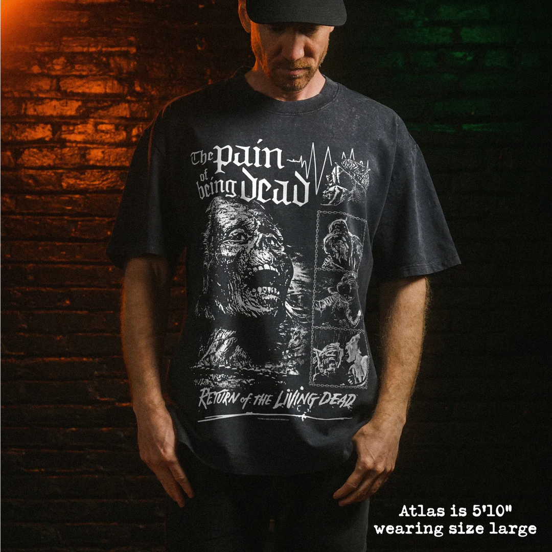 A man wearing a dark stone washed graphic t-shirt with white distressed print that reads 'The pain of being dead' and 'Return of the Living Dead'. He is standing in front of a dark brick wall with dramatic orange and green lighting.