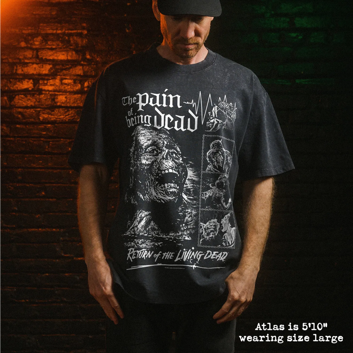 A man wearing a dark stone washed graphic t-shirt with white distressed print that reads 'The pain of being dead' and 'Return of the Living Dead'. He is standing in front of a dark brick wall with dramatic orange and green lighting.