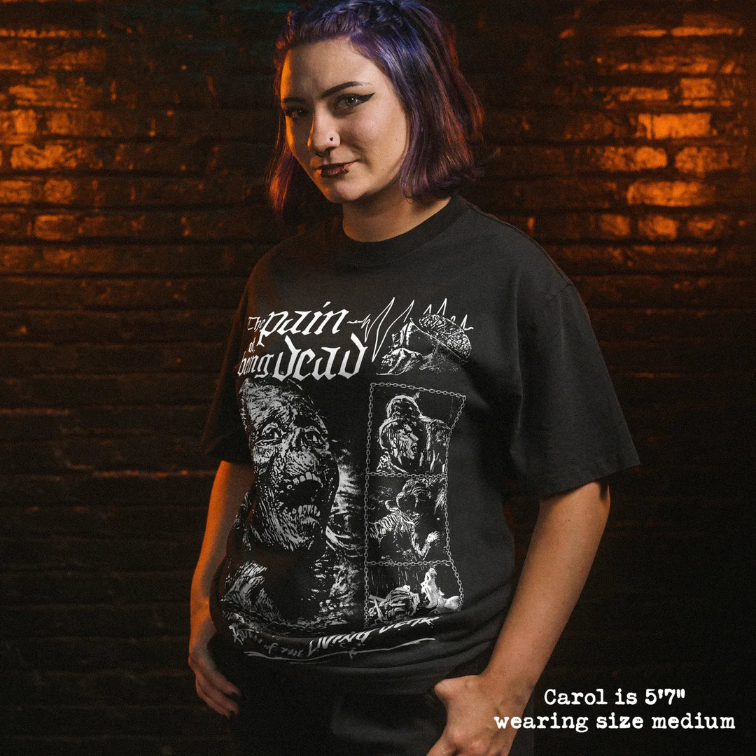A woman with purple hair wearing a black graphic t-shirt with a white print, standing against a dark brick wall with dramatic orange lighting. A text overlay in the bottom right corner reads 'Carol is 5'7" wearing size medium'.