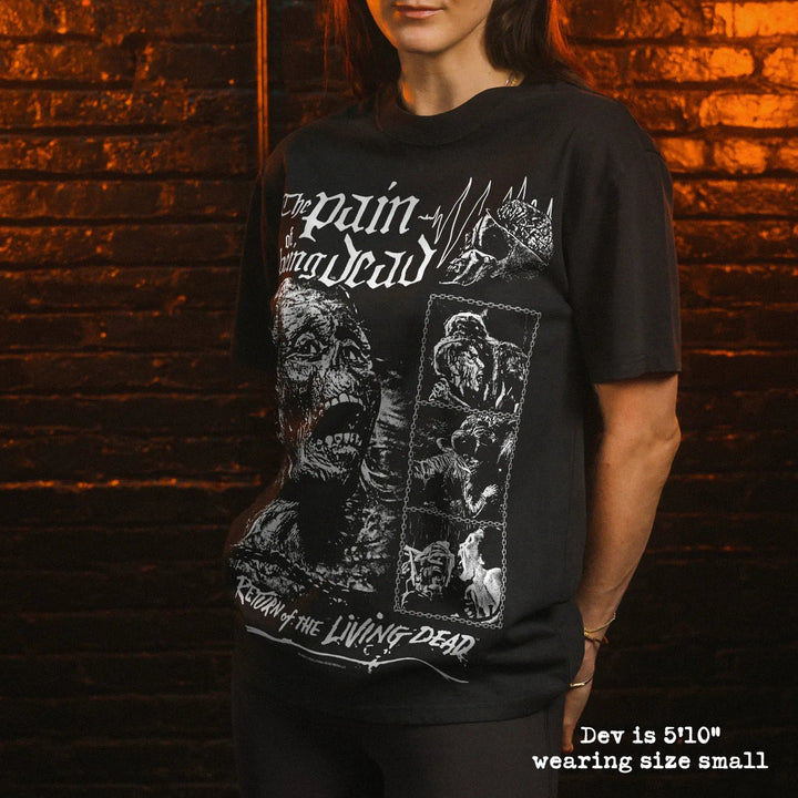 A woman wearing a black graphic t-shirt with white horror-style artwork and text, standing in front of a dark brick wall with warm lighting.