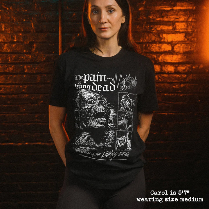 A woman wearing a black graphic t-shirt stands against a warm-lit brick wall. The shirt features a white horror zombie graphic and text.