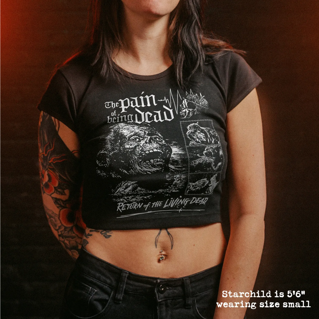 A woman wearing a black short-sleeve graphic crop top with white print reading 'The Pain of Being Dead' and 'Return of the Living Dead,' paired with black jeans against a dark brown background.