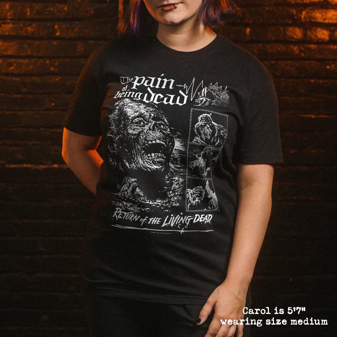 A woman wearing a black graphic t-shirt with white distressed text and illustrations reading 'pain being dead' and 'Return of the Living Dead', posed against a dark brick wall background.