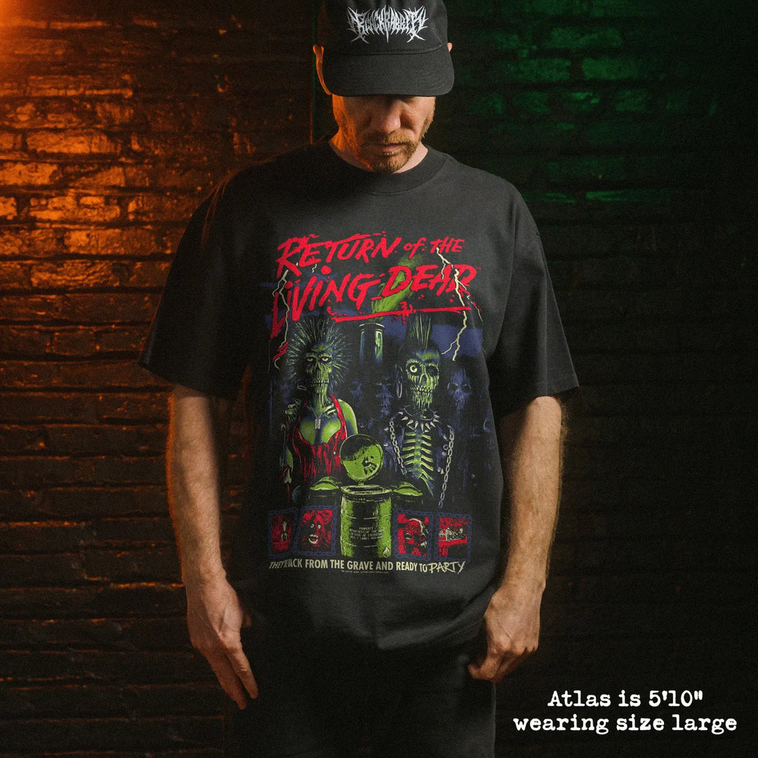A man wearing a black graphic t-shirt with a red and green 'Return of the Living Dead' design, standing against a dark brick wall with dramatic orange and green lighting.