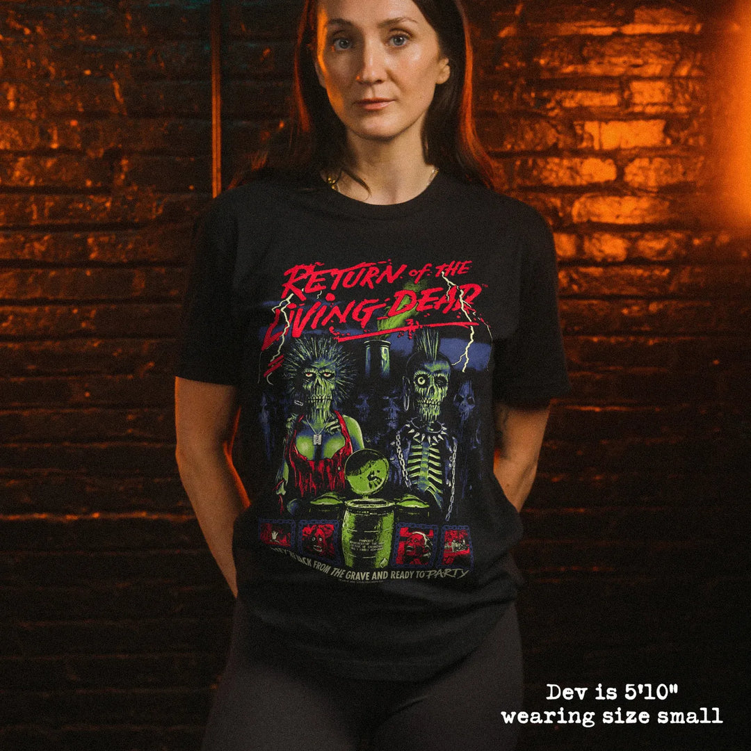 A woman wearing a black graphic t-shirt with a horror-themed print reading 'Return of the Living Dead' and 'They're back from the grave and they're ready to party!'. The background is a dark brick wall with dramatic orange and green lighting.