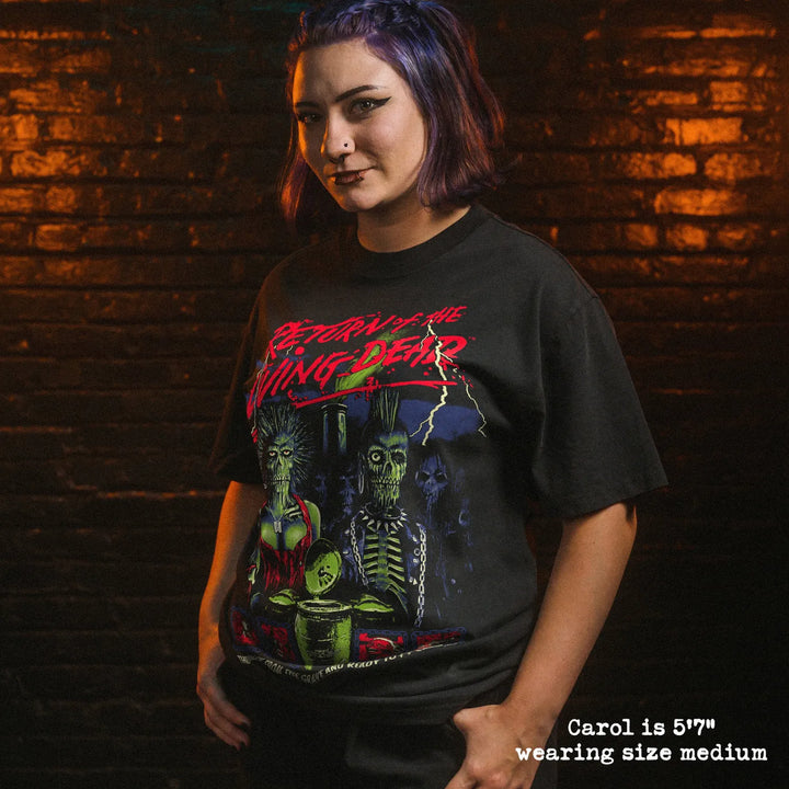 A woman wearing a black graphic t-shirt with a red and green 'Return of the Living Dead' design, standing against a dark brick wall with dramatic orange and green lighting.