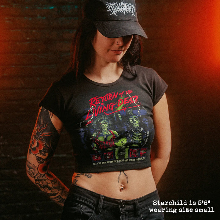 A woman wearing a black graphic crop top t-shirt with 'Return of the Living Dead' text and artwork, paired with a black cap and jeans, set against a dark brick wall with dramatic orange lighting.