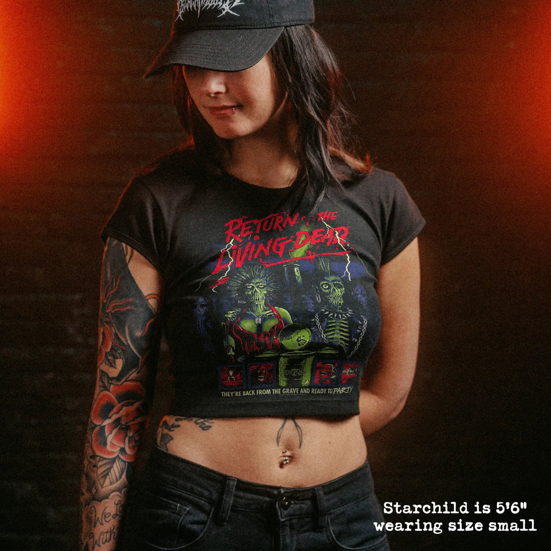 A woman wearing a black graphic crop top t-shirt with 'Return of the Living Dead' text and artwork, paired with a black cap and jeans, set against a dark brick wall with dramatic orange lighting.