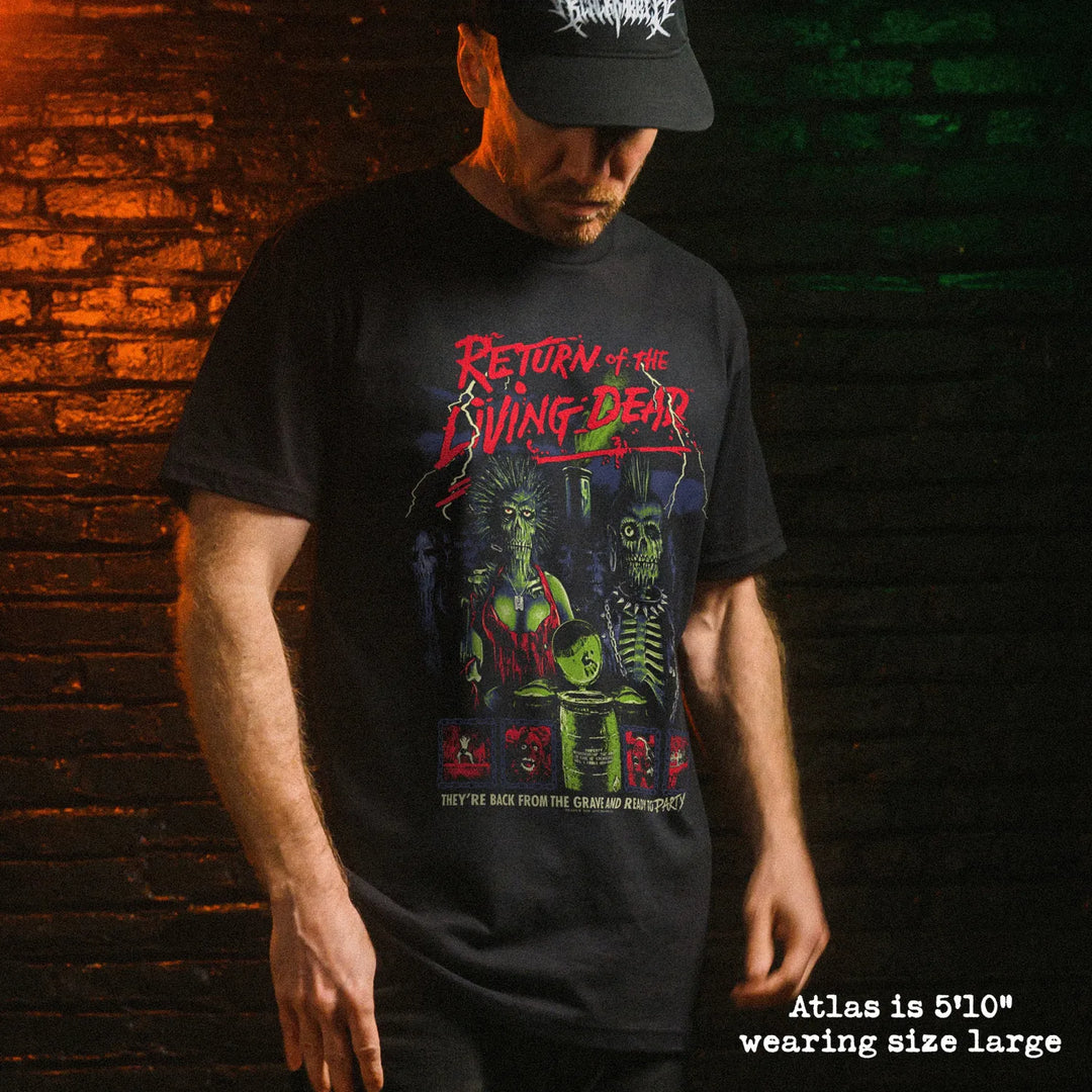 A man wearing a black graphic t-shirt with a horror-themed print reading 'Return of the Living Dead' and 'They're back from the grave and they're ready to party!'. The background is a dark brick wall with dramatic orange and green lighting.