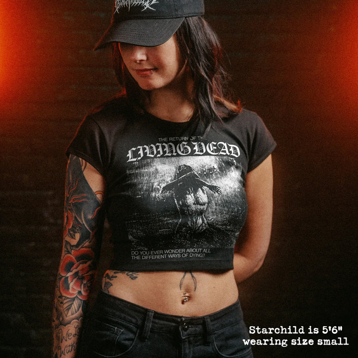 A woman wearing a black crop top t-shirt with a white graphic, set against a dark brick wall with dramatic orange lighting.