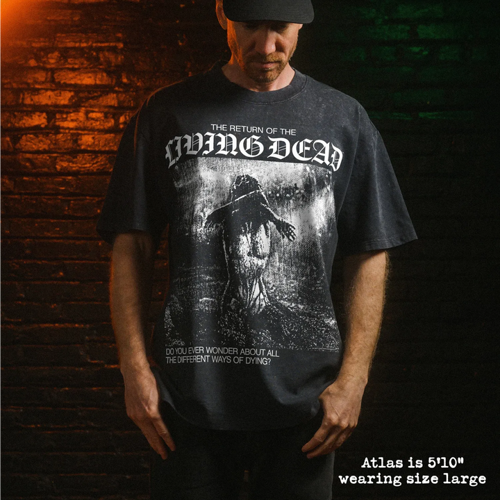 A man wearing a stone-washed graphic t-shirt, standing in front of a dark brick wall with dramatic orange and green lighting. The t-shirt features a white gothic-style logo reading 'The Return of the Living Dead' and a dark, gritty illustration.