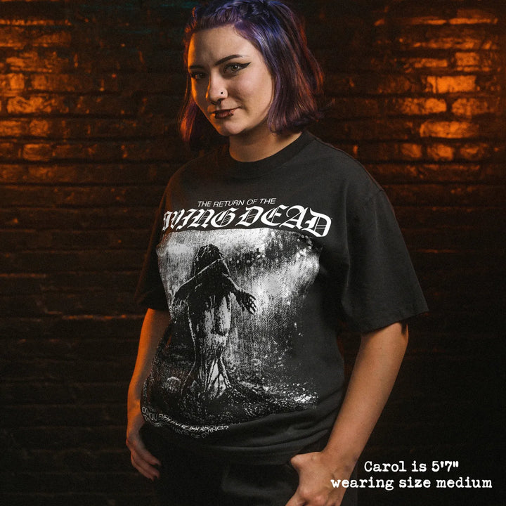 A woman man wearing a black graphic t-shirt, standing in front of a dark brick wall with dramatic orange and green lighting. The t-shirt features a white gothic-style logo reading 'The Return of the Living Dead' and a dark, gritty illustration.
