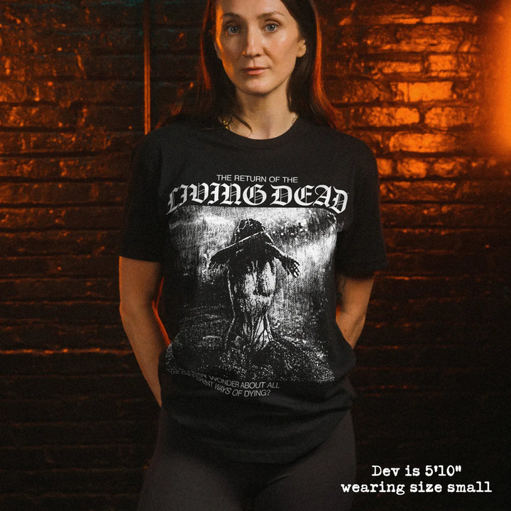 A woman wearing a black graphic t-shirt with the text 'THE RETURN OF THE LIVING DEAD'. She is standing in front of a dark brick wall with warm orange lighting, with text overlay at the bottom right stating 'Dev is 5'10" wearing size small'.