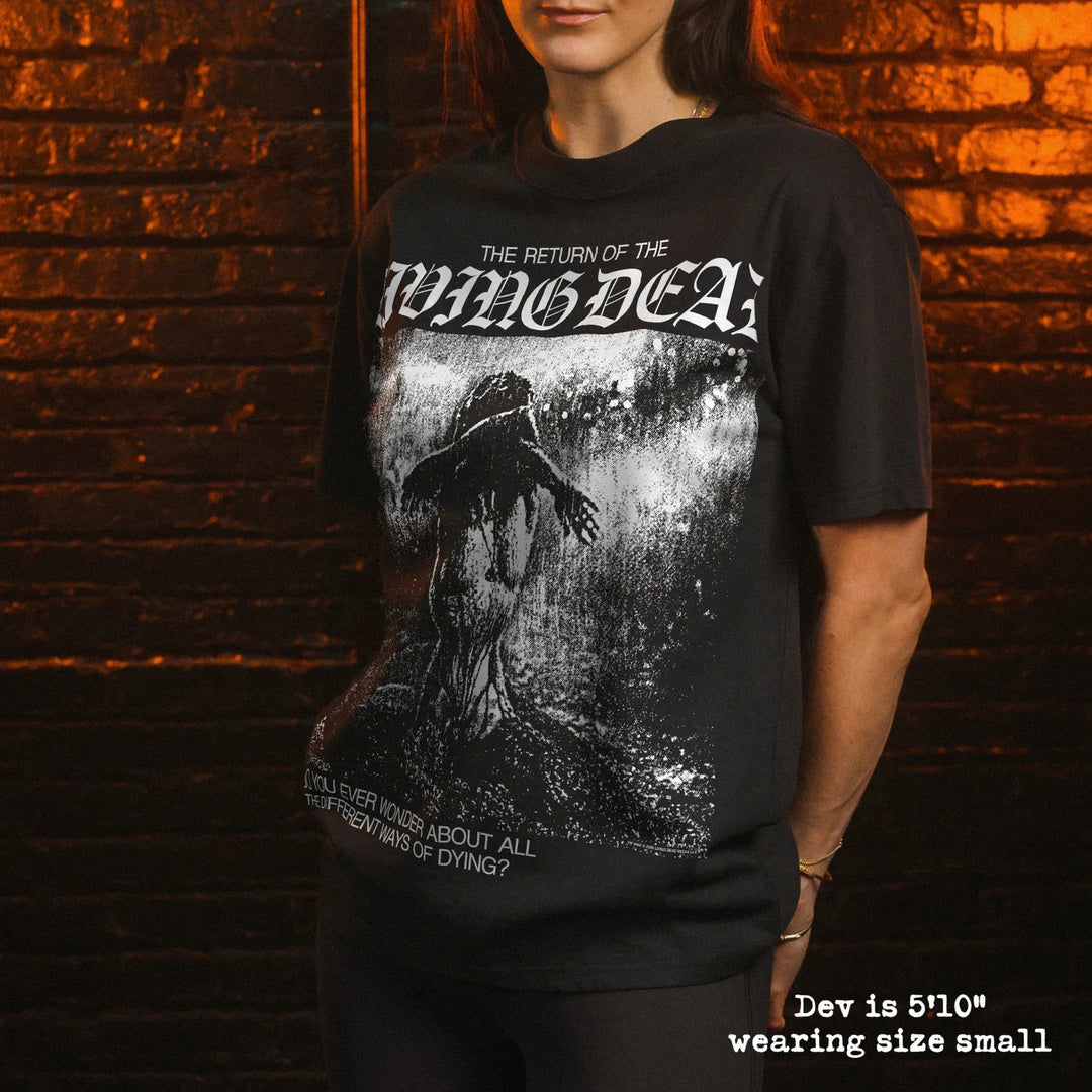 A woman man wearing a black graphic t-shirt, standing in front of a dark brick wall with dramatic orange and green lighting. The t-shirt features a white gothic-style logo reading 'The Return of the Living Dead' and a dark, gritty illustration.