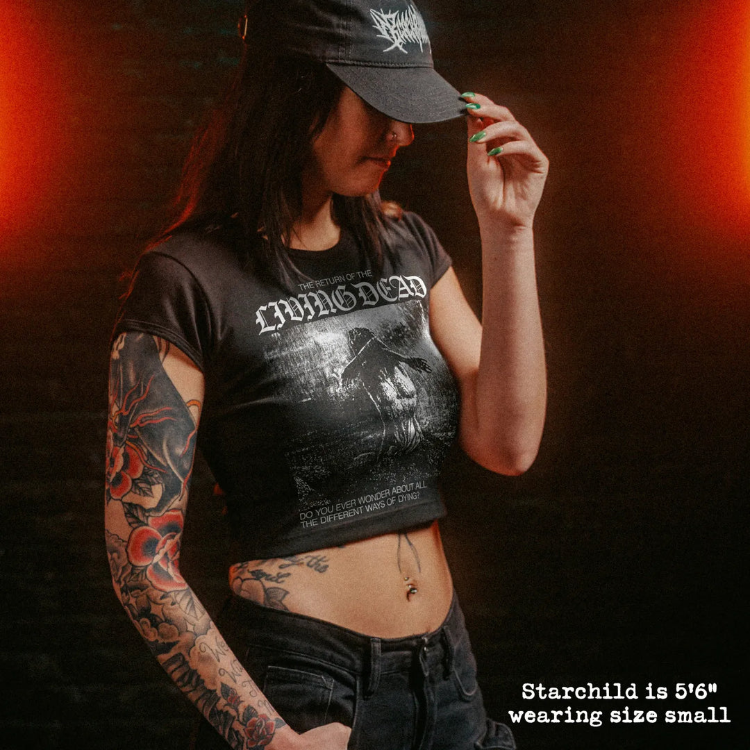 A woman wearing a black crop top t-shirt with a white graphic, set against a dark brick wall with dramatic orange lighting.