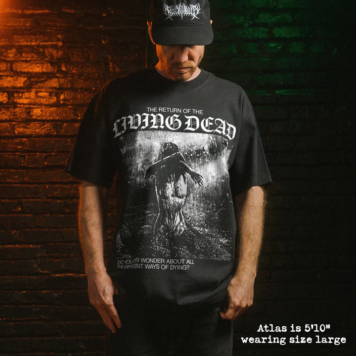 A man wearing a black graphic t-shirt and a black baseball cap, standing in front of a dark brick wall with dramatic orange and green lighting. The t-shirt features a white gothic-style logo reading 'The Return of the Living Dead' and a dark, gritty illustration.