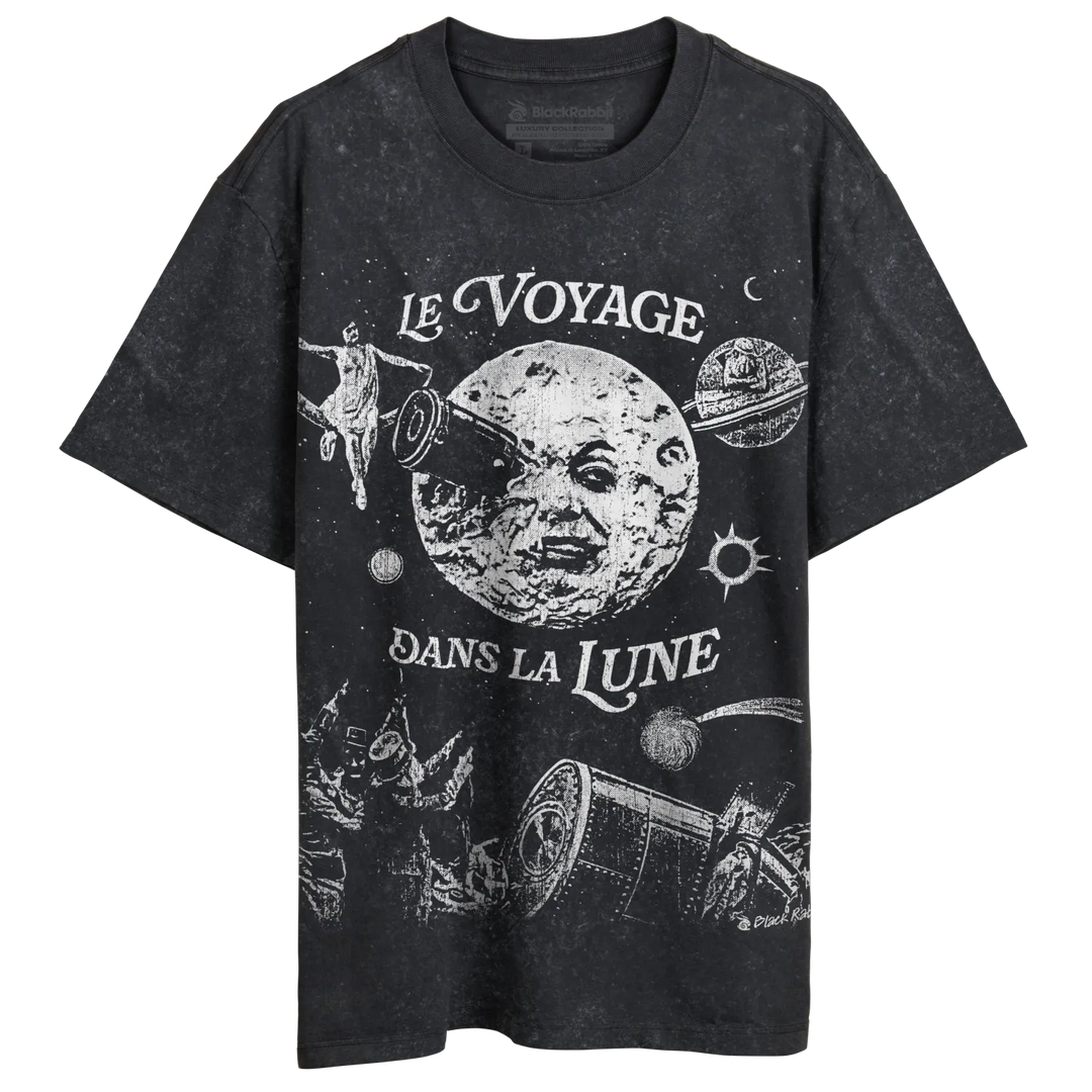 A faded black t-shirt with a vintage-style white graphic depicting a man on the moon and other space-themed imagery, along with the French phrase ’LE VOYAGE DANS LA LUNE’ (A Trip to the Moon).