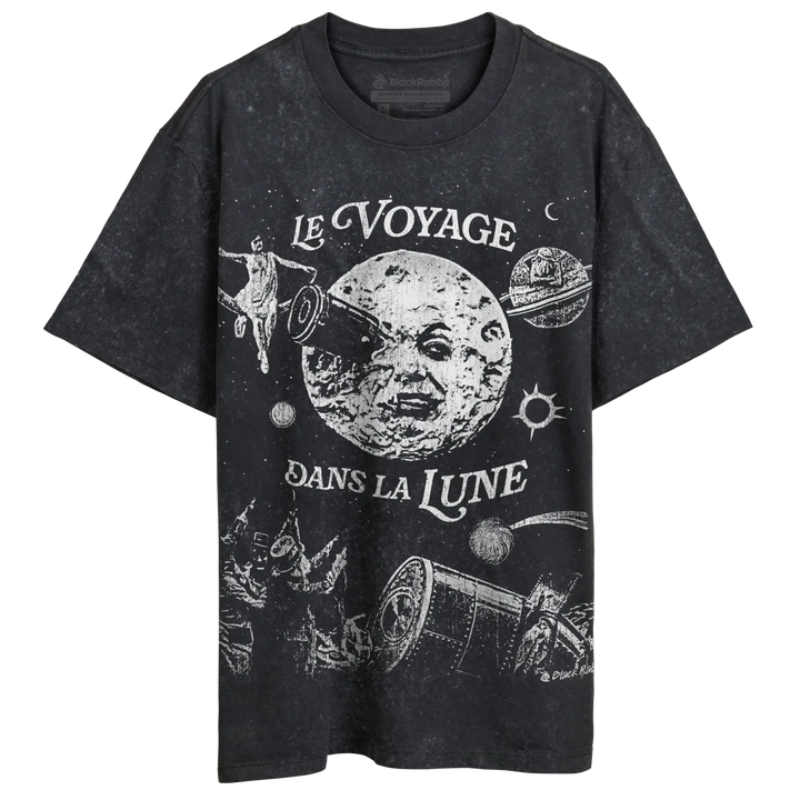 A faded black t-shirt with a vintage-style white graphic depicting a man on the moon and other space-themed imagery, along with the French phrase ’LE VOYAGE DANS LA LUNE’ (A Trip to the Moon).